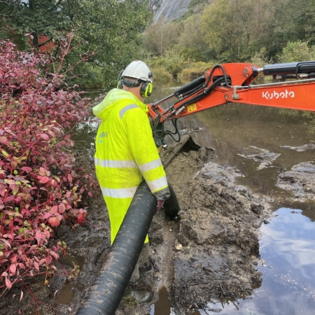 Arbeider i verneutstyr bruker rør ved elveutgraving med gravemaskin.