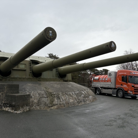 Oransje lastebil ved en gammel kanoninstallasjon under grå himmel.