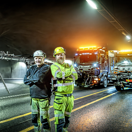 To arbeidere i gule uniformer foran lastebiler i en tunnel om natten.