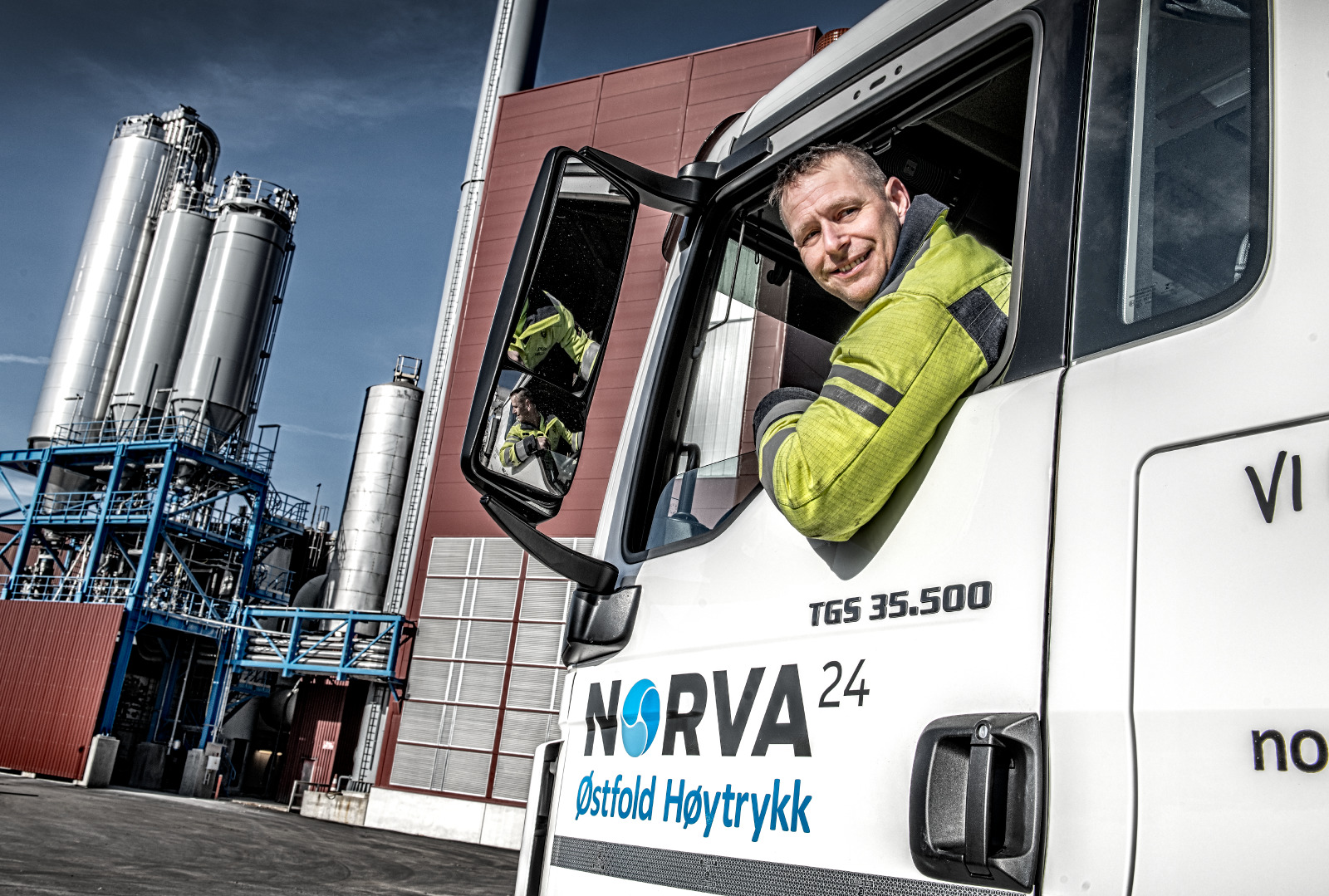 Smilende Norv24-sjåfør i refleksutstyr lener seg ut av lastebilførerhuset på industriområde