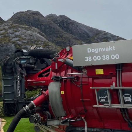 En stor rød sugebil med slanger og skilt for døgnvakt foran fjell.
