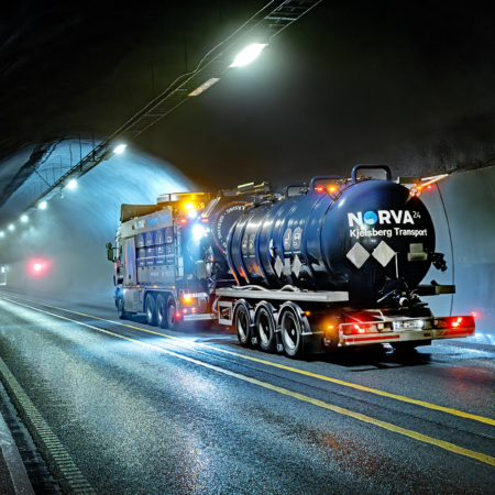 Svart lastebil med "Norva Kjelsberg Transport" i en opplyst tunnel.
