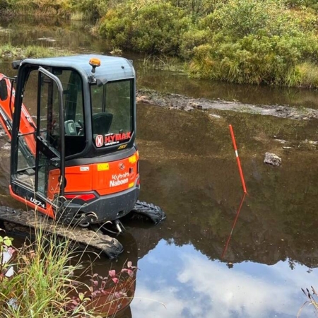 En gravemaskin arbeider i en sumpete elv med rød markeringsstolpe.