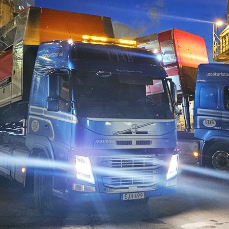 Två blå lastbilar som lossar på en industriplats under natten, upplysta av starka strålkastare och anläggningsljus.