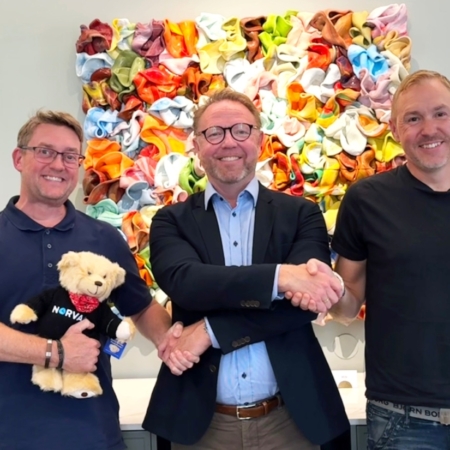 Tre leende män skakar hand framför färgglada konstverk och firar ett affärspartnerskap.