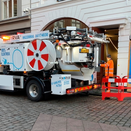 Elektrisk Norva24-servicebil med slangar står vid en byggnad, arbetare i orange dräkt.