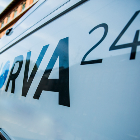 Närbild av Norva24-logotypen på sidan av en vit servicebil med en suddig stadsbakgrund.