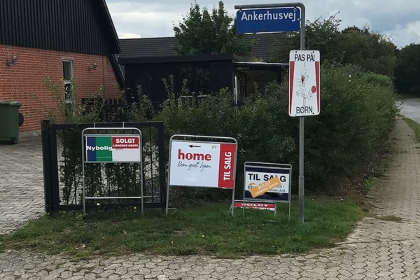 Bostadshörn med försäljningsskyltar, en "se upp för barn"-skylt och ett rött tegelhus i bakgrunden.
