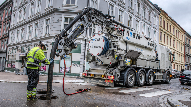 Two workers inspecting pipes using equipment near an open service van on a city street.