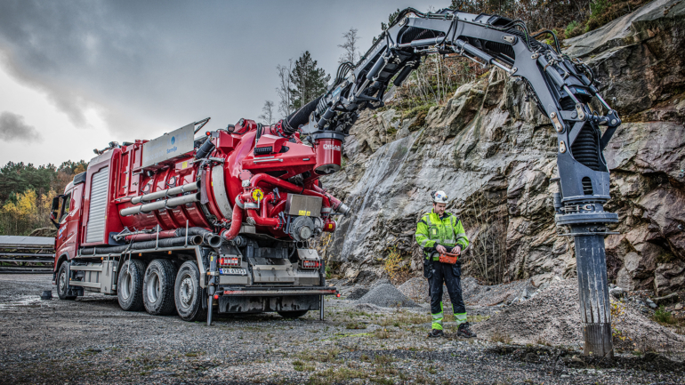 Operator using a red vacuum truck with a large excavator arm for industrial cleaning near rocky terrain.
