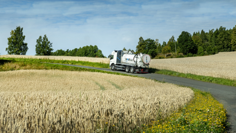 A Norva24 truck drives along a rural road surrounded by golden wheat fields under a bright blue sky.