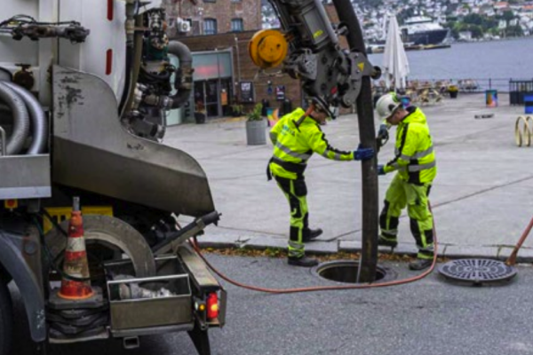 Two Norva24 workers in yellow uniforms performing sewer maintenance in an urban area by the waterfront.