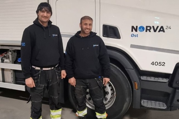 Two Norva24 employees in black hoodies smiling, standing beside a company truck indoors.