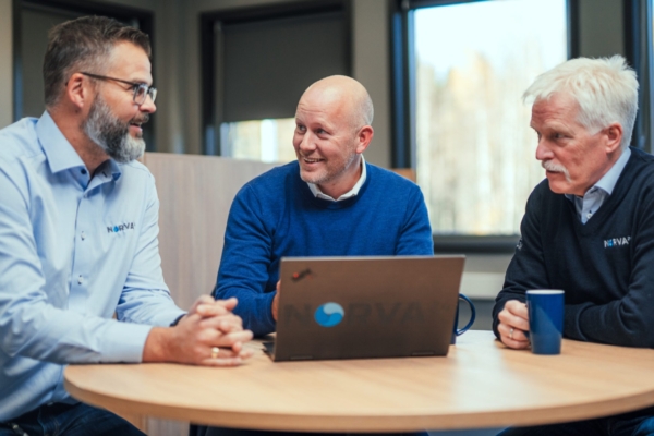 Three Norva24 employees in a meeting, smiling and discussing around a table with a laptop.