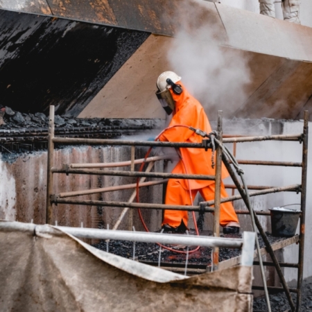 Worker in protective gear cleaning an industrial structure using a high-pressure water jet amidst rising steam.