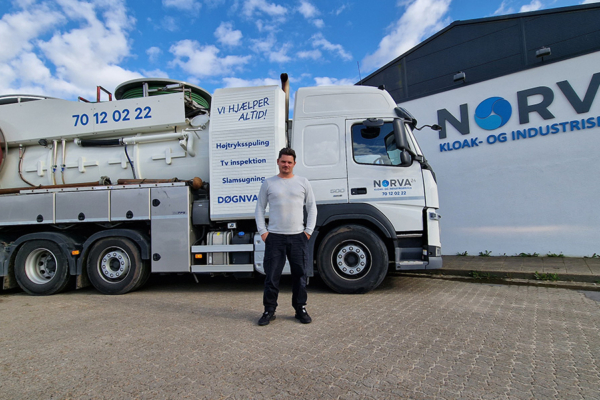 Man stands by a Norva24 truck with branding, parked outside the company building on a sunny day.