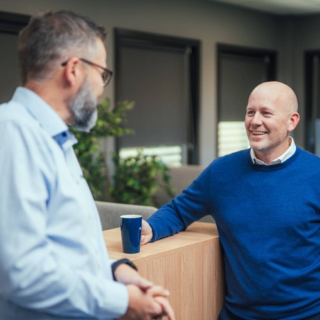 Two men in an office having a conversation, one leaning on a wooden desk with a coffee cup.