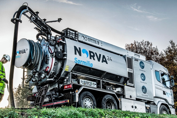 Worker operating Norva24 Sørmiljø vacuum truck on grassy roadside.