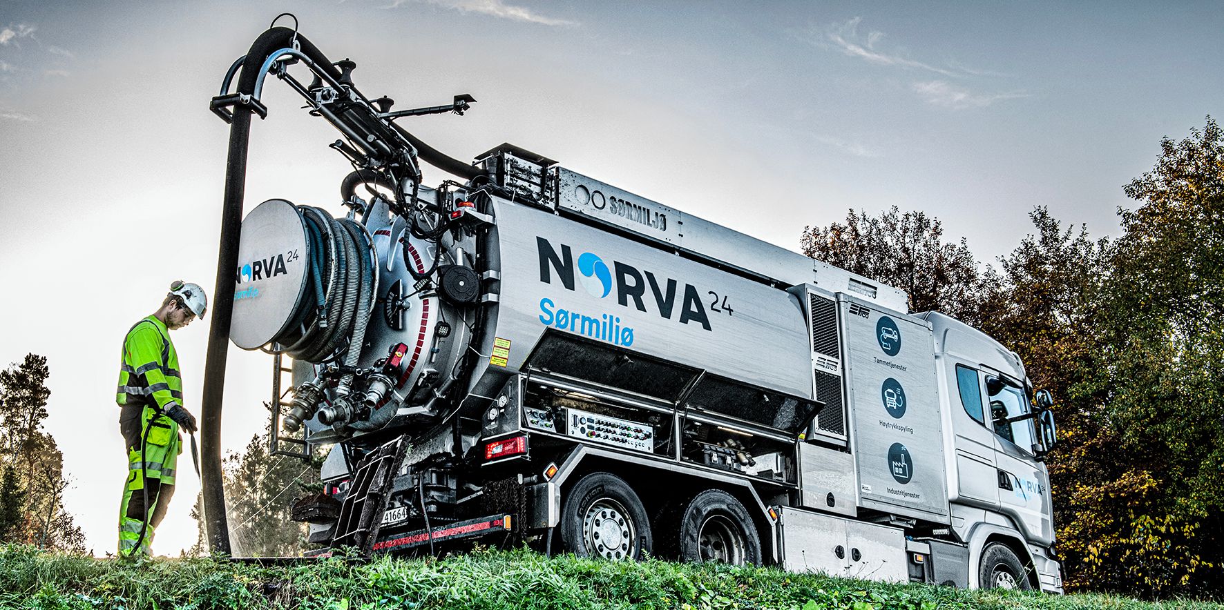 Worker operating Norva24 Sørmiljø vacuum truck on grassy roadside.