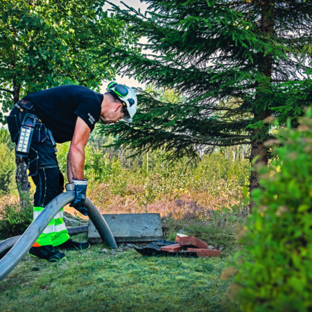 Arbeider støvsuger en septiktank utendørs med verneutstyr i et skogkledd område