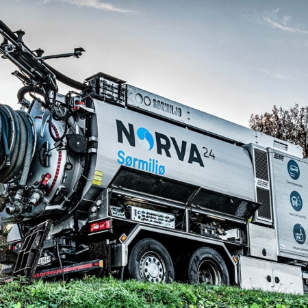 Worker operating Norva24 Sørmiljø vacuum truck on grassy roadside.