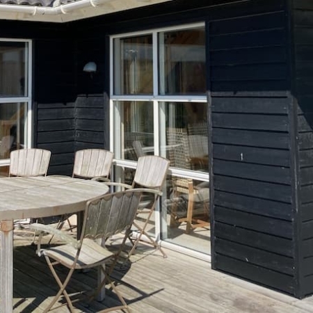 Wooden patio with a round table and chairs outside a modern black summer house under a blue sky.